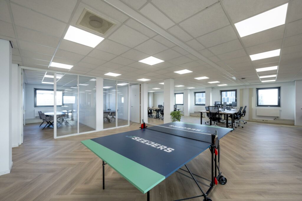 Modern kantoorpand aan de Joan Muyskenweg met glazen vergaderruimtes, werkplekken en een tafeltennistafel van Seeders op de voorgrond.